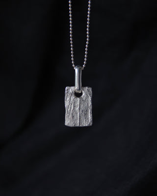 A textured silver rectangular pendant necklace (tag pendant) hanging from a silver ball chain against a dark black fabric background.