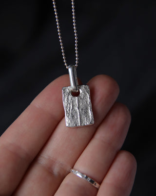 A silver textured pendant necklace held on a person's fingers against a dark background. The pendant is a rough, rectangular silver tag with a bail and a delicate silver ball chain.