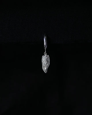 A single silver huggie hoop earring with a textured, irregularly shaped dangling charm, shown against a solid black background.